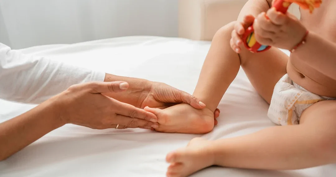 A woman gently holds a baby's foot while the child sits on a bed, creating a tender moment of connection.
