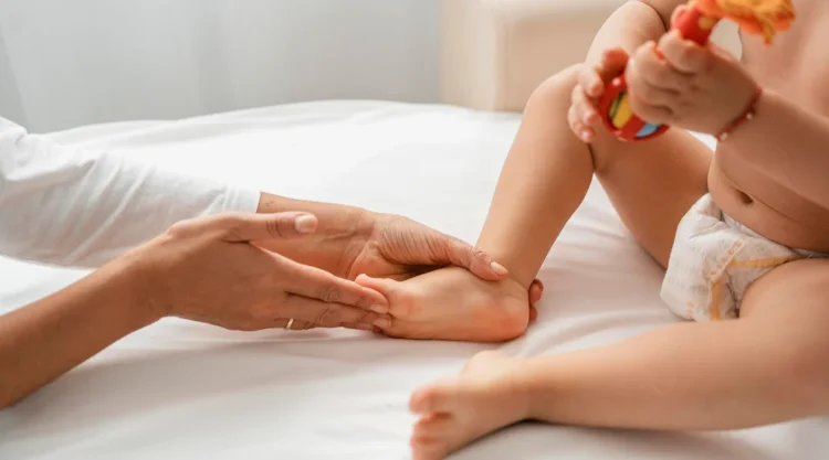 A woman gently holds a baby's foot while the child sits on a bed, creating a tender moment of connection.