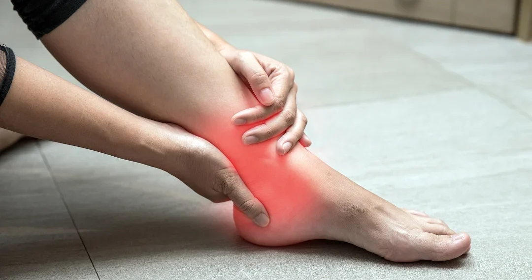 A person sitting on the ground, holding their painful ankle with a pained expression on their face.